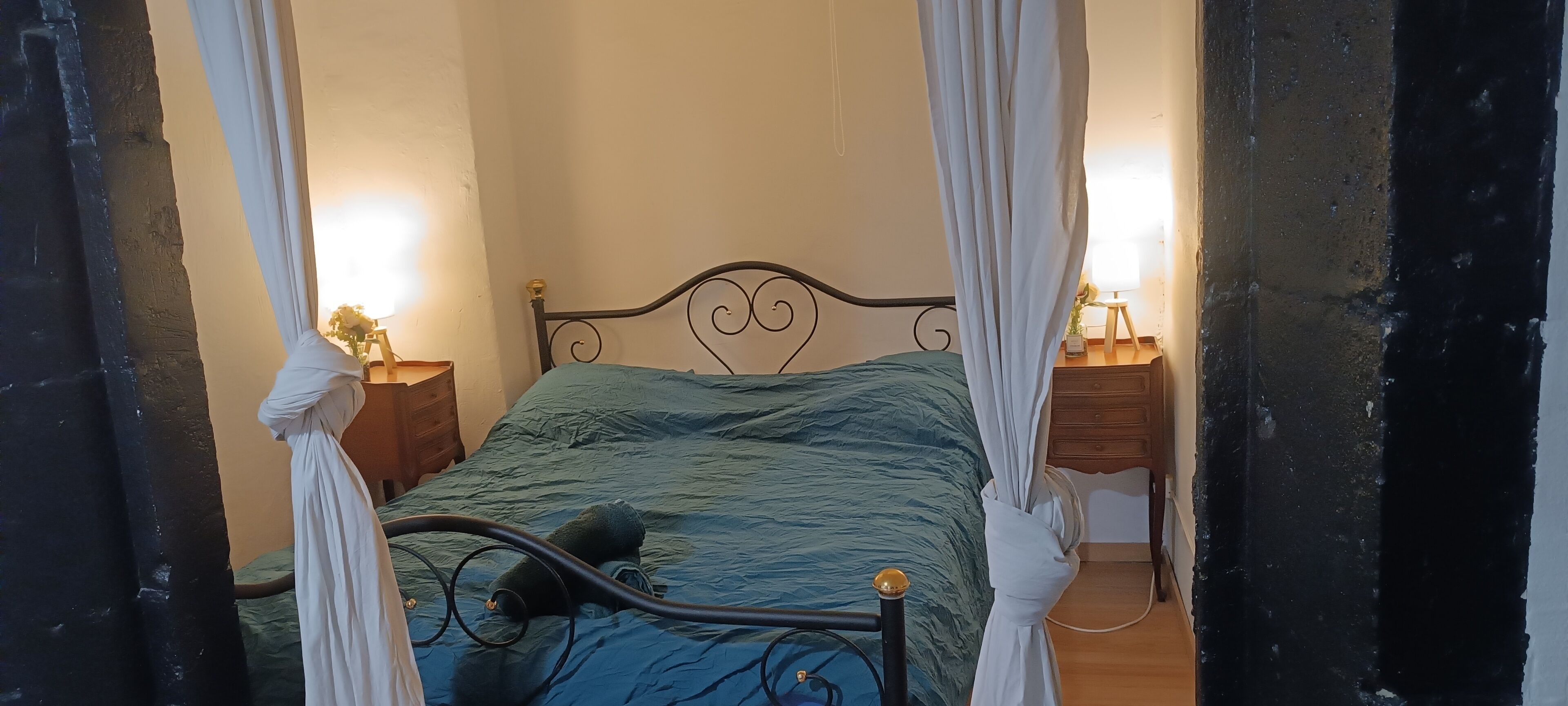 A bed with a metal frame, a blue bedspread, and white curtains. There are two bedside tables with lamps.
