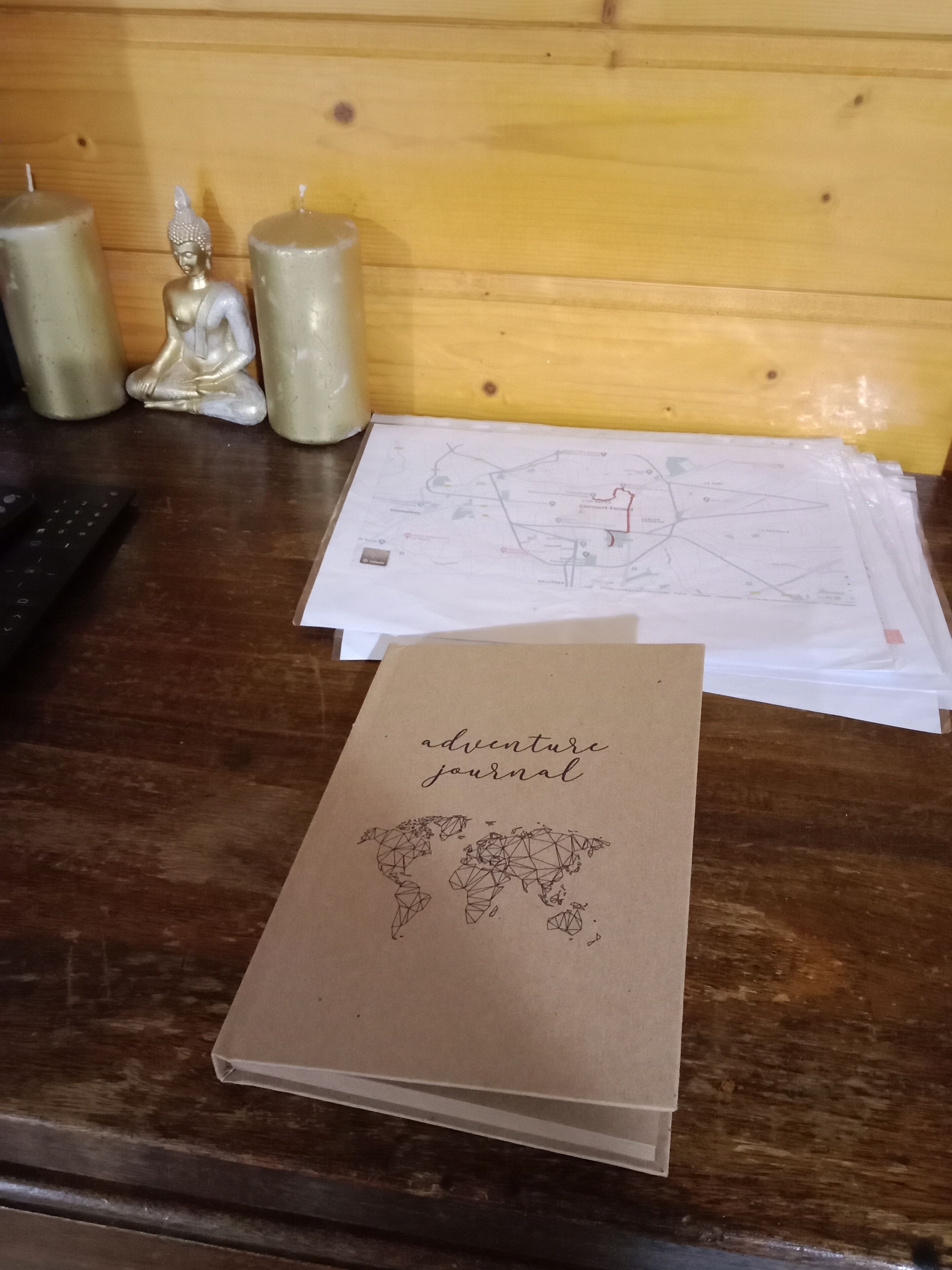 Un escritorio de madera con un cuaderno marrón titulado “diario de aventuras”, una ilustración de un mapa mundial y un mapa con contorno rojo.