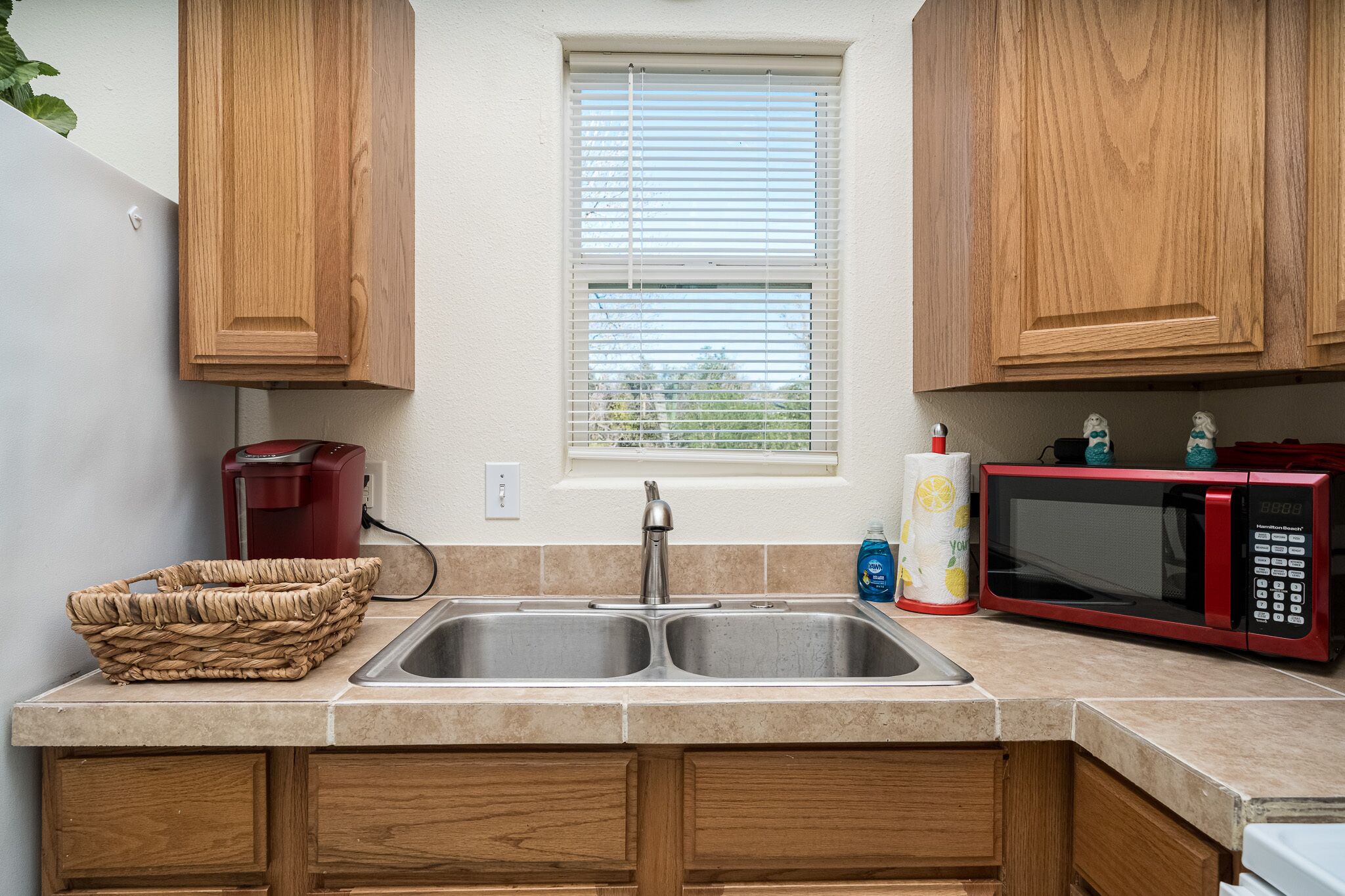 Una cocina con armarios de madera, un lavamanos doble, una cafetera roja, un microondas y una cesta.