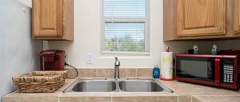 Una cocina con armarios de madera, un lavamanos doble, una cafetera roja, un microondas y una cesta.