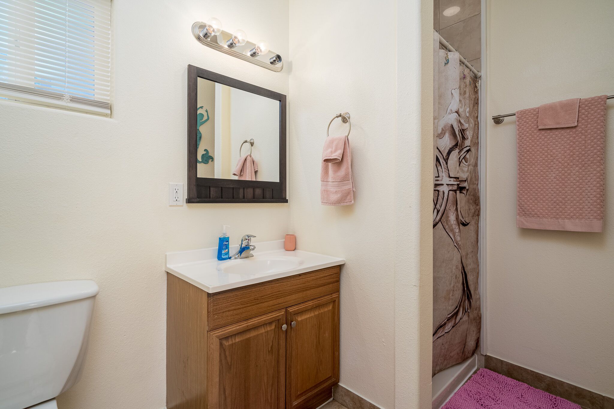 A bathroom with a white sink, wooden vanity, a mirror with lights, a towel rack with pink towels, and a shower with a pink mat.