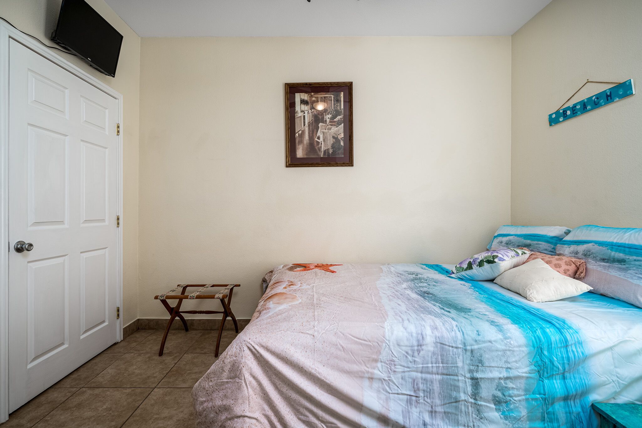 A bedroom with a bed, a small wooden stool, a framed picture on the wall, and a 'BEACH' sign.