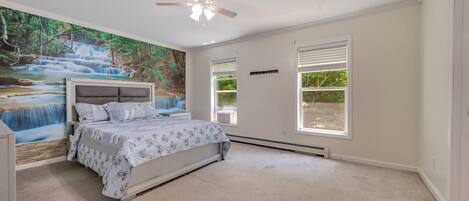 Un dormitorio con cama, ventilador de techo, un mural y ventanas amplias.