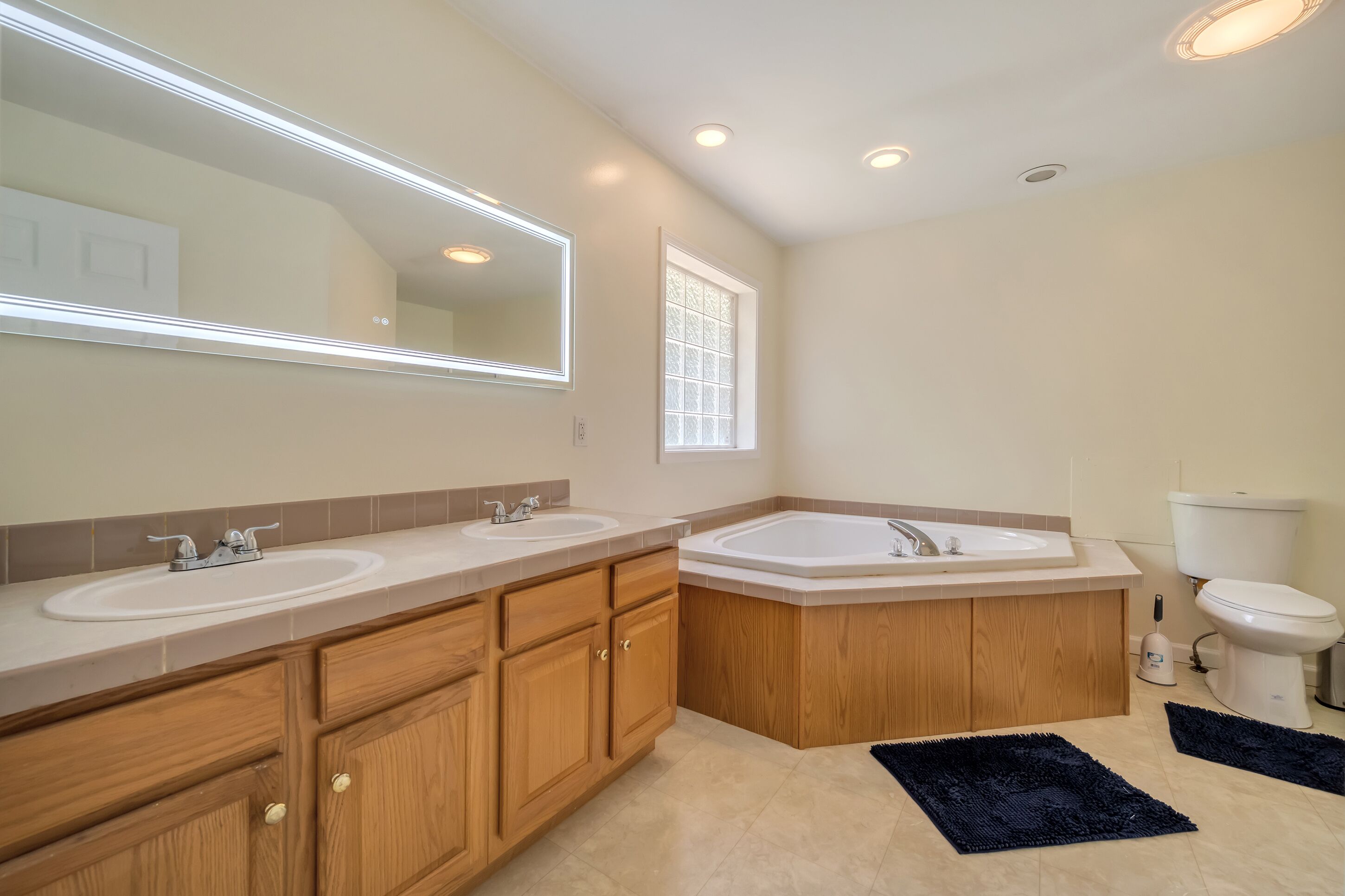 A modern bathroom with two sinks, a bathtub, wooden cabinets, a large mirror, recessed ceiling lights, and a window.