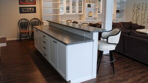 A modern kitchen with white cabinetry and a granite countertop. There are two chairs and a sofa in the living area.