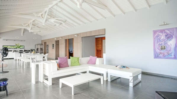 A spacious living area with a white sectional sofa, a dining table, and a kitchen in the background.