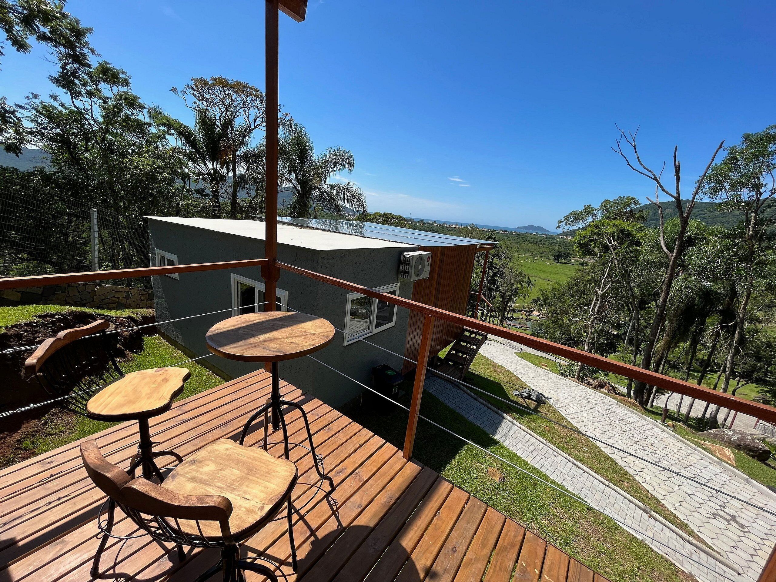 Une terrasse en bois avec deux tables et des chaises, d’où l’on peut admirer une petite maison dotée d’un toit en métal et d’un mur en pierre.