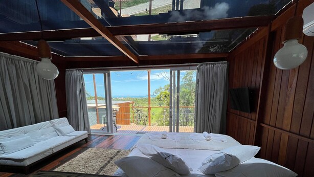 A bedroom with a large bed, a sofa, and a dining table with fruit. - Casa de Vidro - Hospedar com Propósito (Florianópolis)