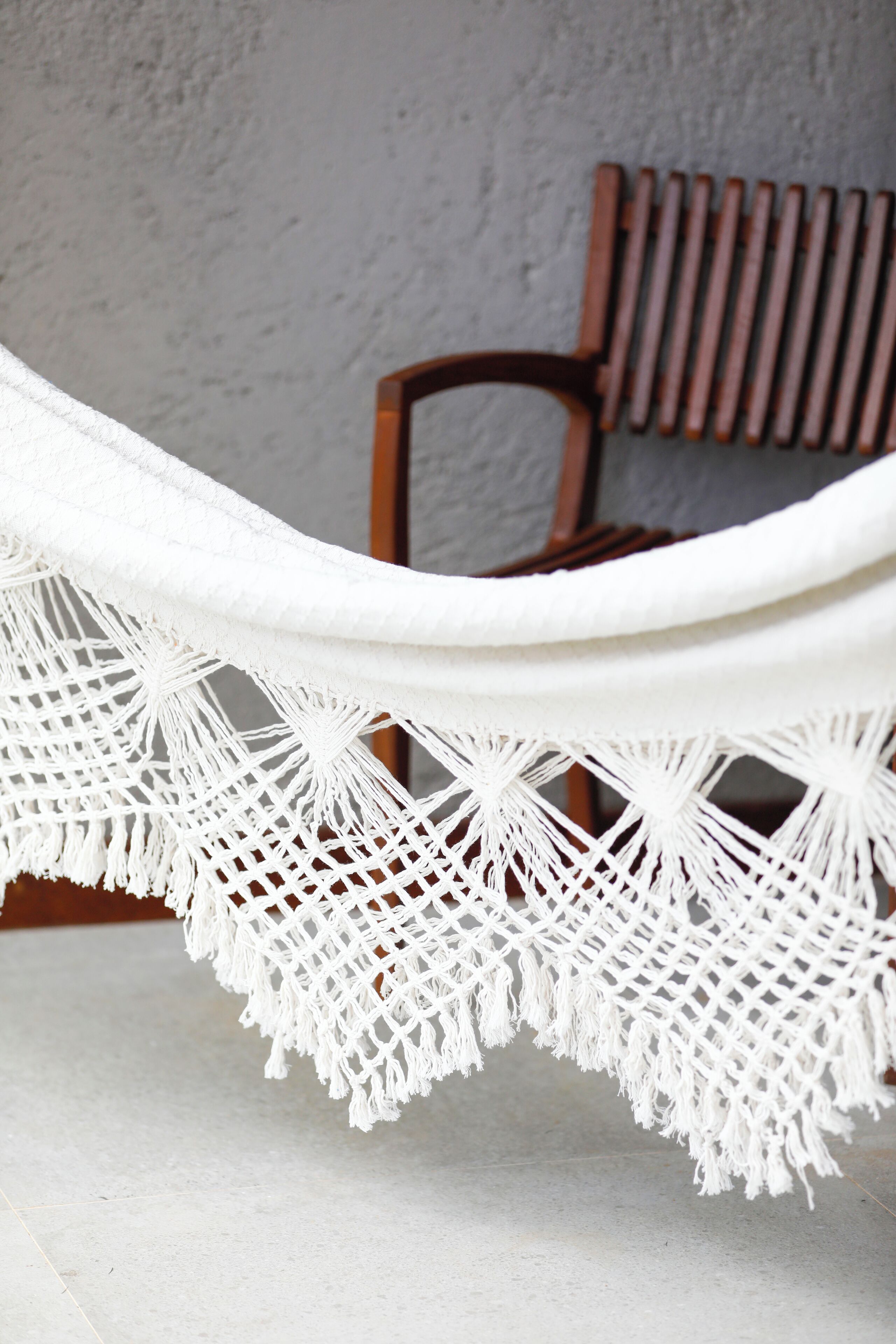 Un hamac orné d’une bordure en macramé blanc, une chaise en bois et un mur gris avec un radiateur.