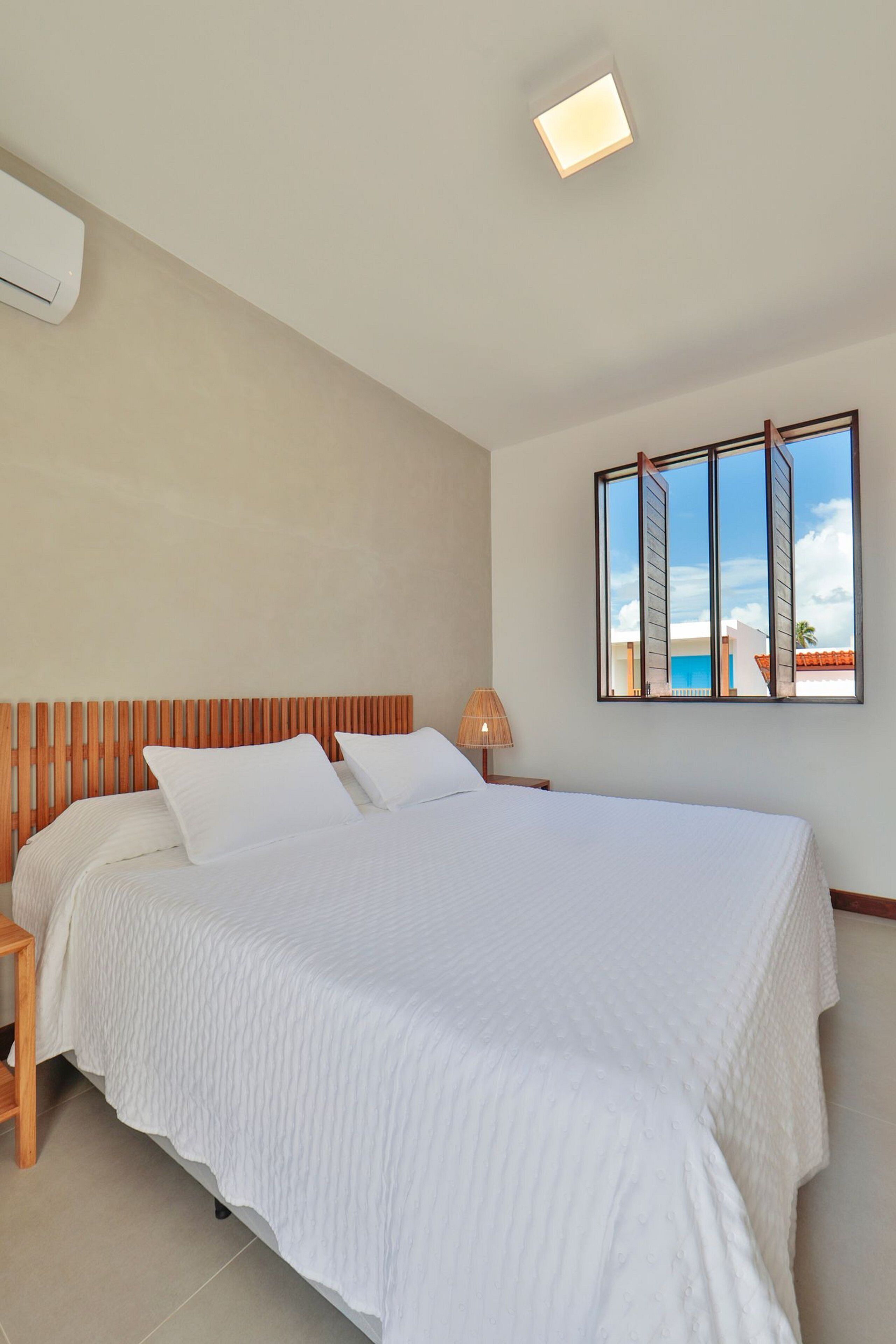 A bedroom with a large bed, a wooden headboard, a bedside lamp, and a window with a view of buildings and a blue sky.