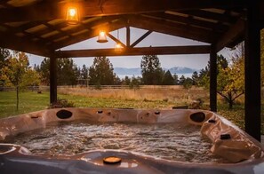 Relax in a serene hot tub under a shaded pergola, surrounded by a lush grassy field and majestic mountains.