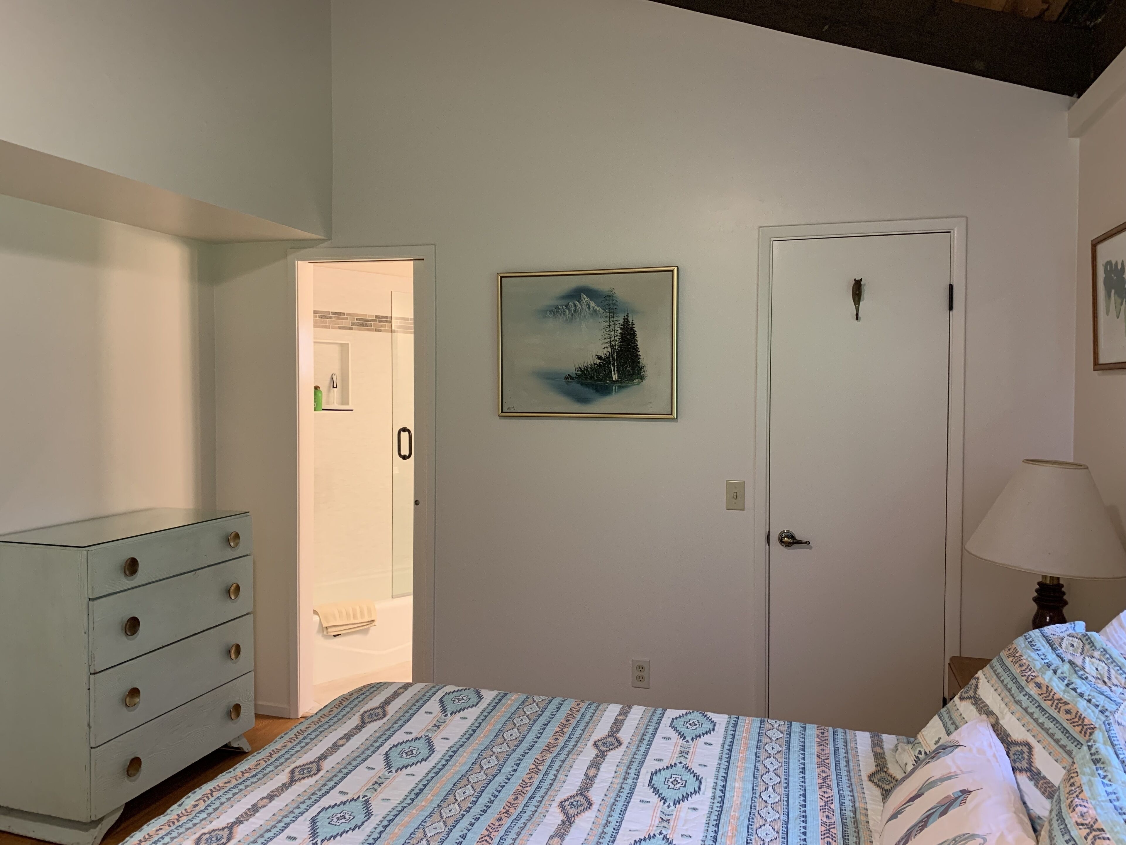 Une chambre à coucher avec un lit, une commode, une porte et un tableau encadré au mur.