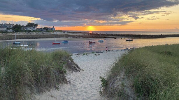 Una playa con barcos, un atardecer y casas al fondo.