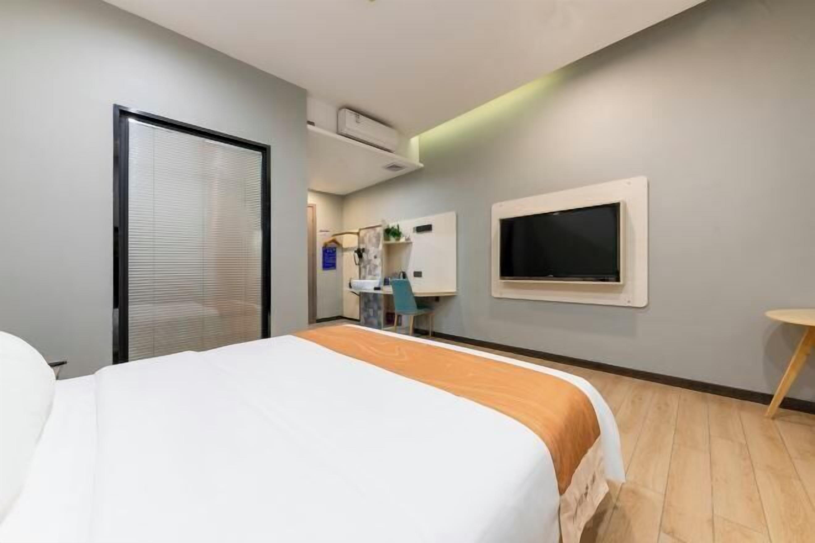 A hotel room with a bed, a desk, a chair, a TV, and an air conditioning unit.