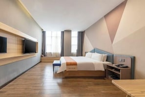 Ein Hotelzimmer mit Bett, einem an der Wand montierten Fernseher, einem Schreibtisch und einem Fenster mit Vorhängen.