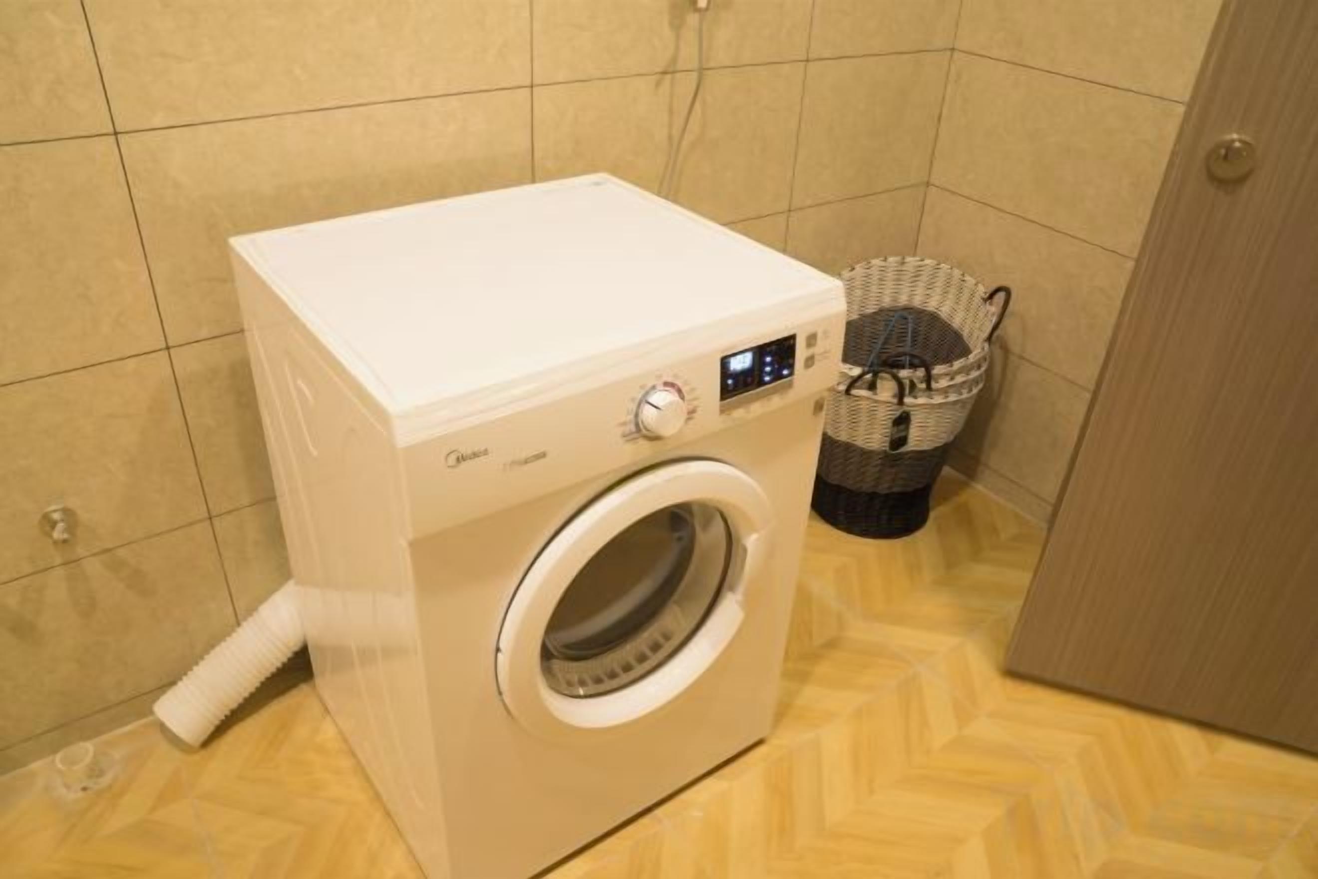 Un lave-linge moderne et compact, doté d’un écran numérique, placé dans un coin d’une pièce dont le sol est recouvert d’un parquet à motifs en chevrons et dont les murs sont carrelés.