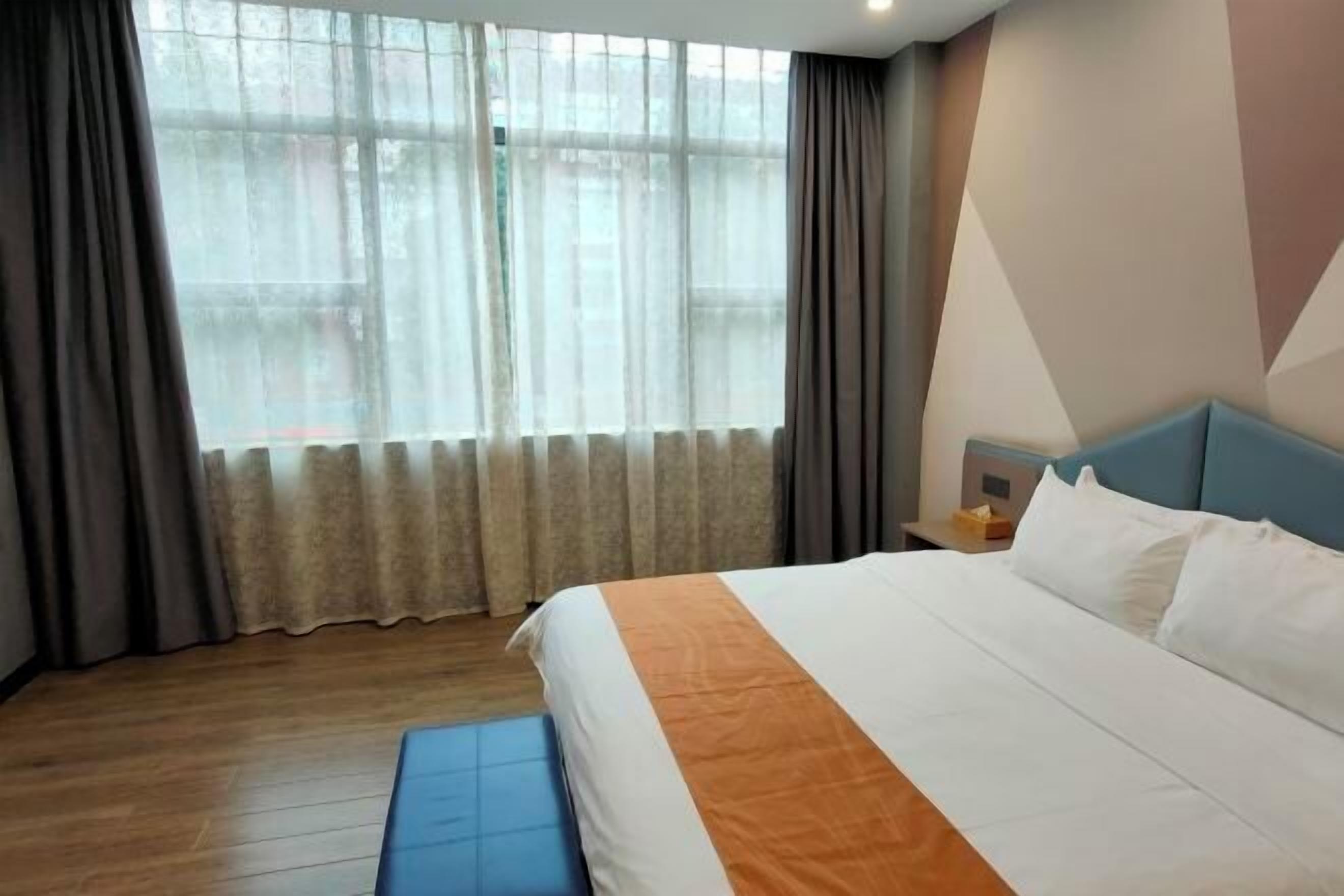 A hotel room with a large bed, a wooden floor, a window with sheer curtains, and a geometric wall design.