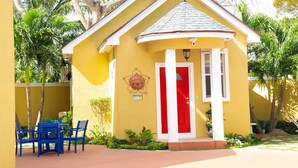Un edificio amarillo con puerta roja, columnas blancas y un pequeño patio con sillas azules.