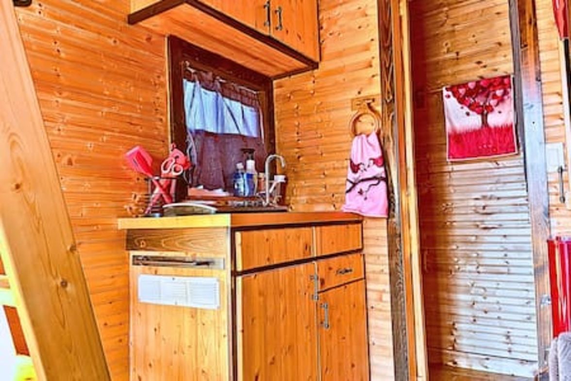 L’intérieur d’un chalet en bois comprend un coin cuisine avec des placards, un évier et une fenêtre avec des rideaux.