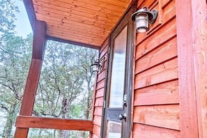 A wooden exterior with a door and window, a porch with a roof, and a view of trees.