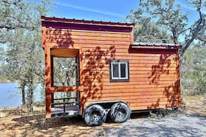 A small wooden cabin with a single window and a door, a flat roof, and a trailer attached.