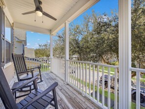 A covered porch with two rocking chairs, a ceiling fan, and a view of a residential street and trees.