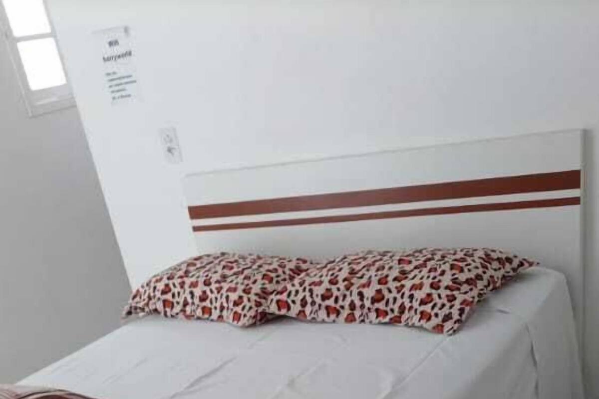 A single bed with a white sheet and a patterned blanket, a headboard with a brown stripe, and a tiled floor.