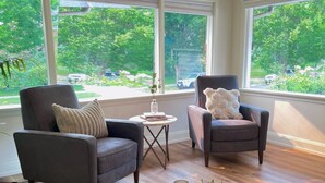 Un salon moderne avec deux fauteuils gris, une petite table basse et une vue sur un paysage verdoyant grâce à de grandes fenêtres.