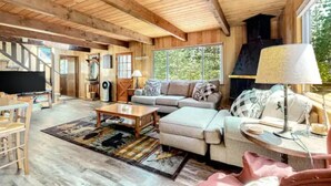 A cozy cabin interior with a wooden ceiling, a large window, a sofa, a coffee table, and a TV.