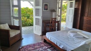 Un dormitorio con cama, una silla, un escritorio y vistas a la vegetación a través de contraventanas abiertas.