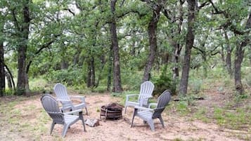 A forested area with two Adirondack chairs and a small fire pit.