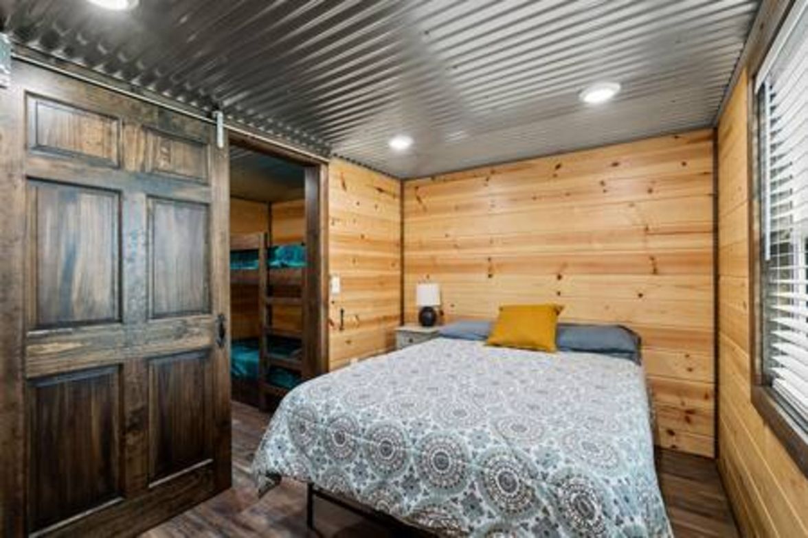 Un dormitorio con techo de madera, una cama con colcha estampada, una mesita de noche con lámpara y una ventana con persianas.