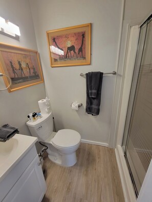 A bathroom with a toilet, sink, and a shower area. There are framed pictures on the wall and a towel rack with a towel hanging on it. - Book one week or more and get 10% discount
Stylish 2 BR Suite in West Seattle!
 (Seattle)