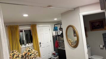 A bedroom with a bed, a window with curtains, a door, a mirror, and a bathroom with a toilet and sink.