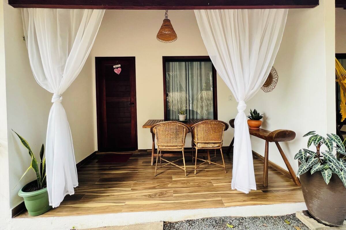Un espacio al aire libre cubierto con sillas de mimbre, una mesa, cortinas blancas y plantas en macetas.