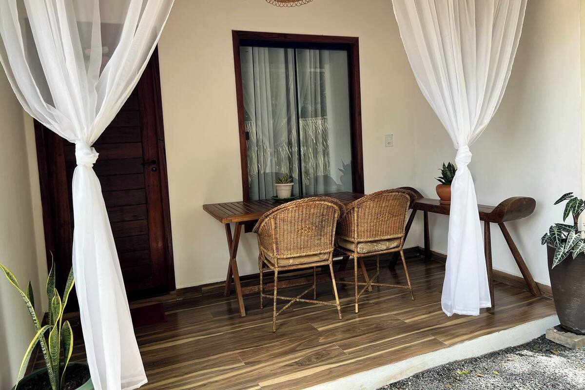 Un porche con piso de madera, dos sillas de mimbre, una mesa y una planta en maceta.
