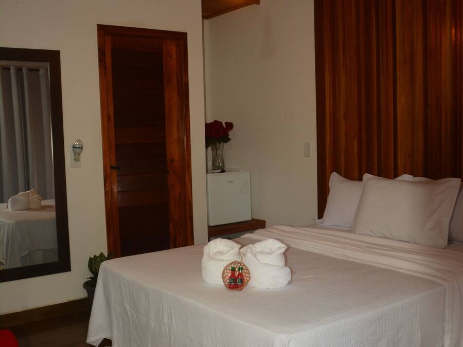 Una cama bien hecha con sábanas blancas, techo de madera y una silla roja en una habitación con piso de madera.