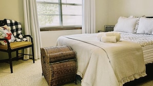 Une chambre à coucher avec un lit, une chaise, une commode en osier et une fenêtre avec des stores.