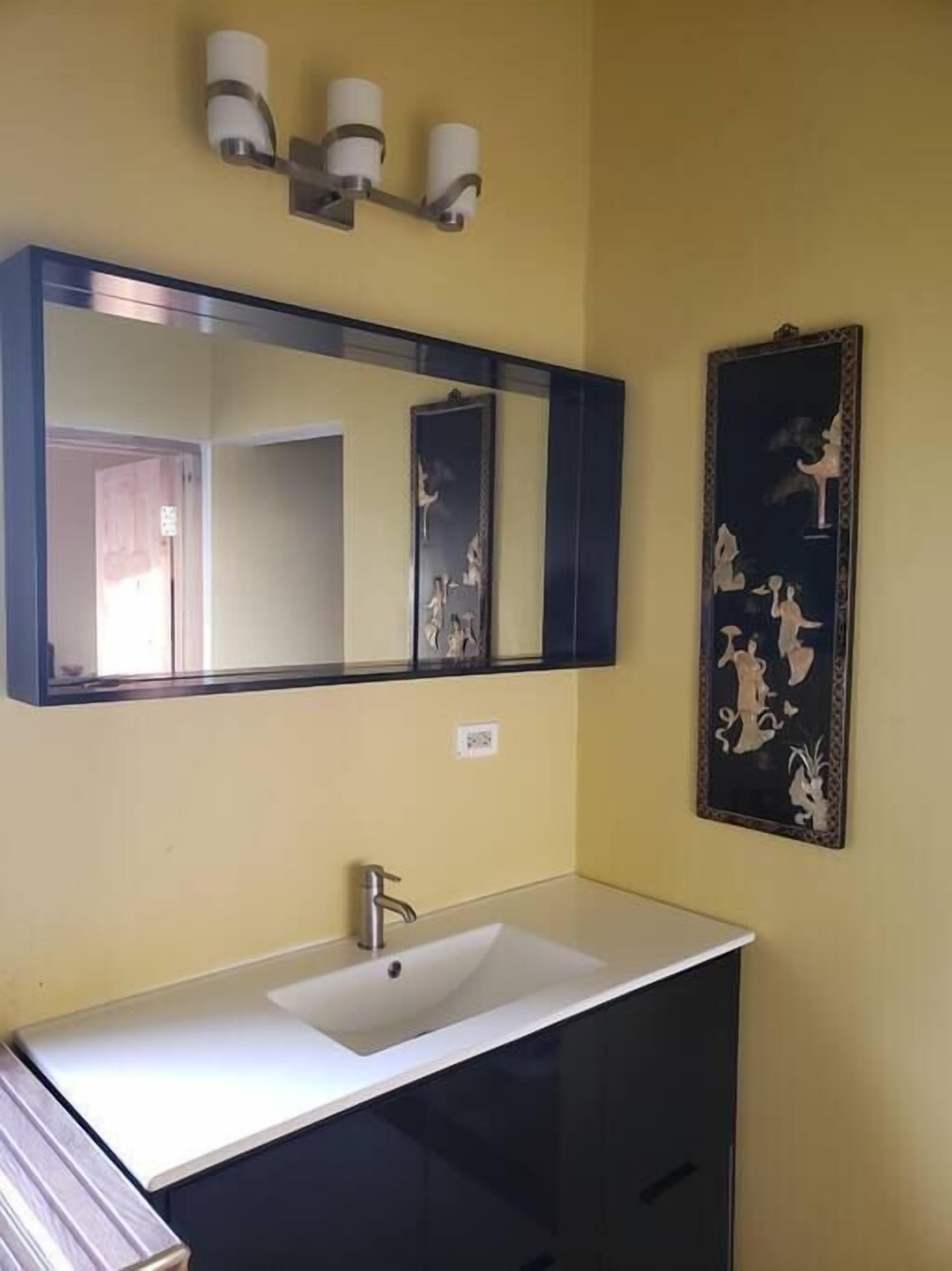 Une salle de bain avec un lavabo blanc, des meubles sombres, un grand miroir et un luminaire fixé au mur.