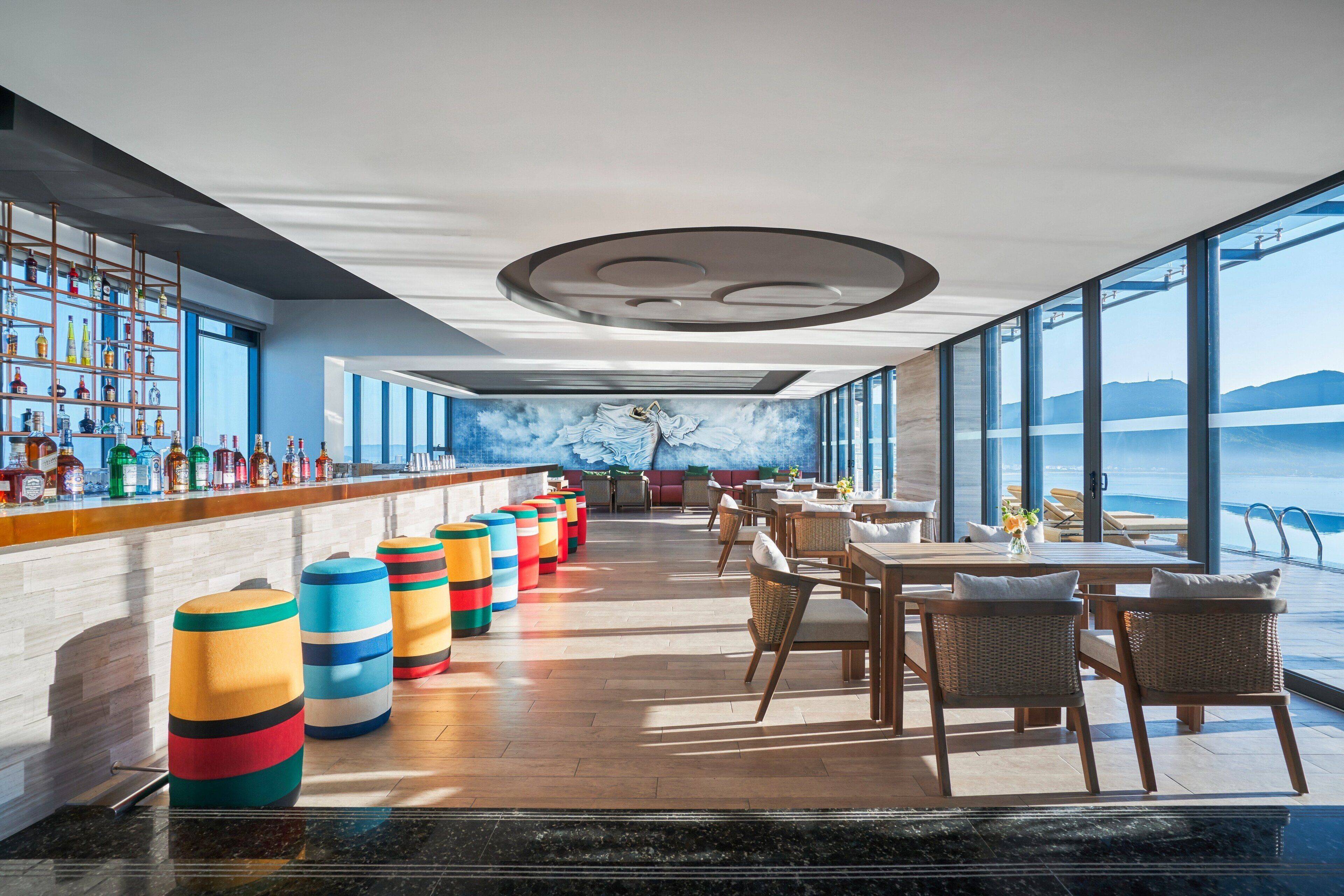 Un espace bar moderne avec un long comptoir, des tabourets colorés et une vue sur la mer grâce à de grandes fenêtres.