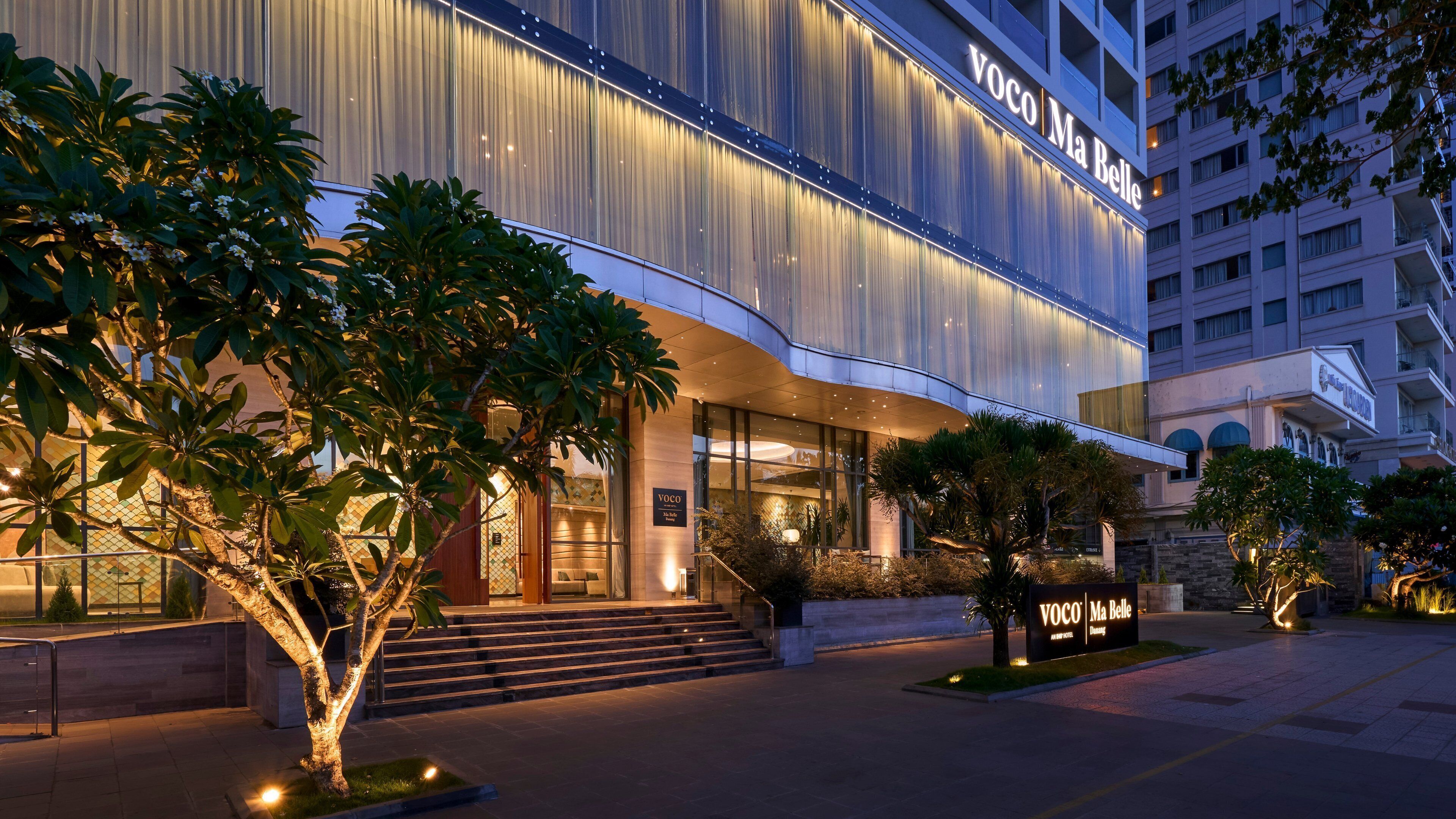 A modern hotel entrance with a large sign reading "VOCO Ma Belle" and a staircase leading up to the entrance.