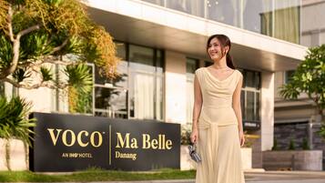 Una mujer de pie frente al letrero de un hotel que muestra el nombre "VOCO Ma Belle" y "UN HOTEL IHG".