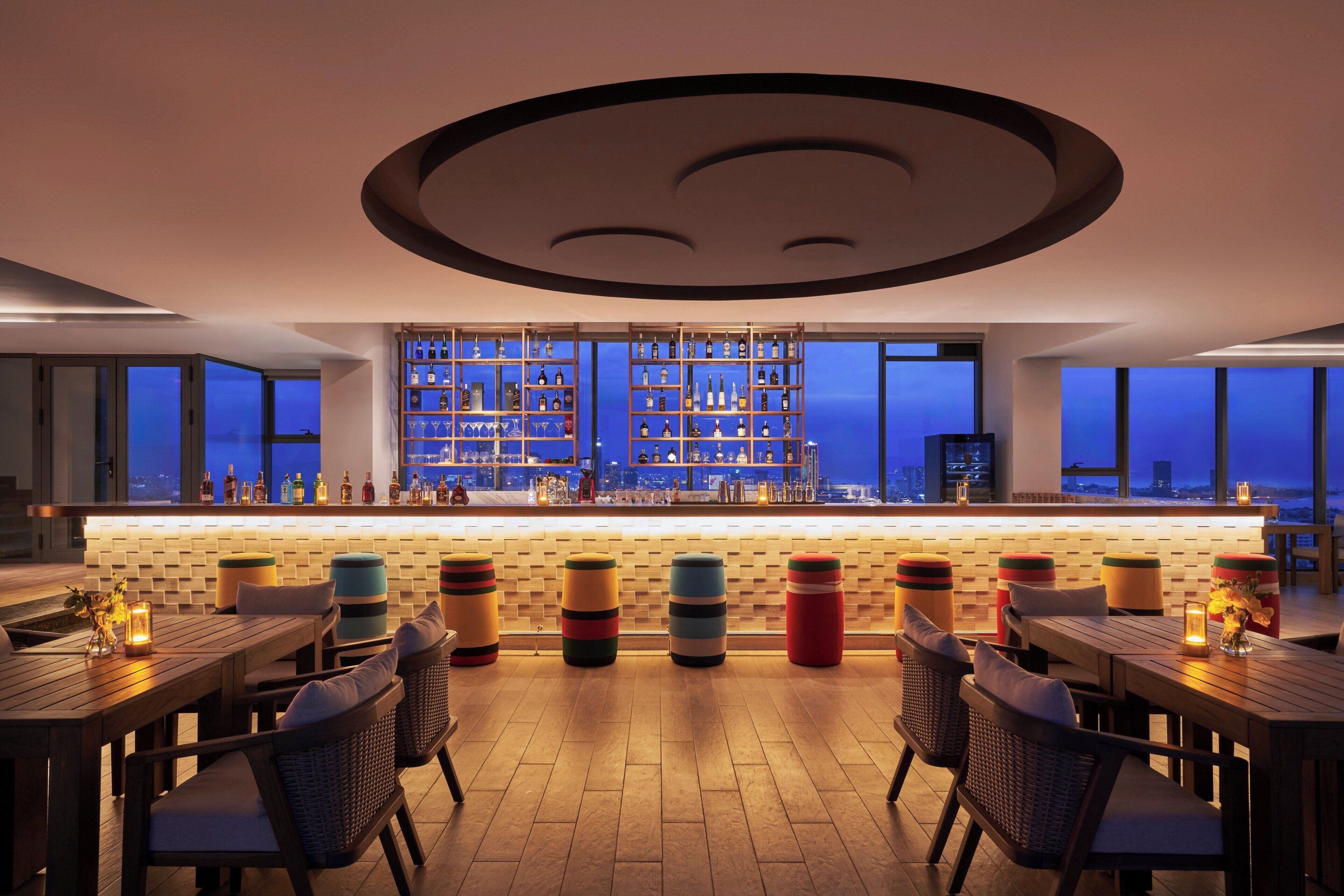 A bar area with a city view, wooden furniture, and a variety of bottles and glasses on shelves.