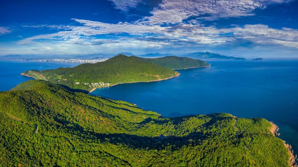 A coastal landscape with a small island, dense green forest, and a clear blue sea.