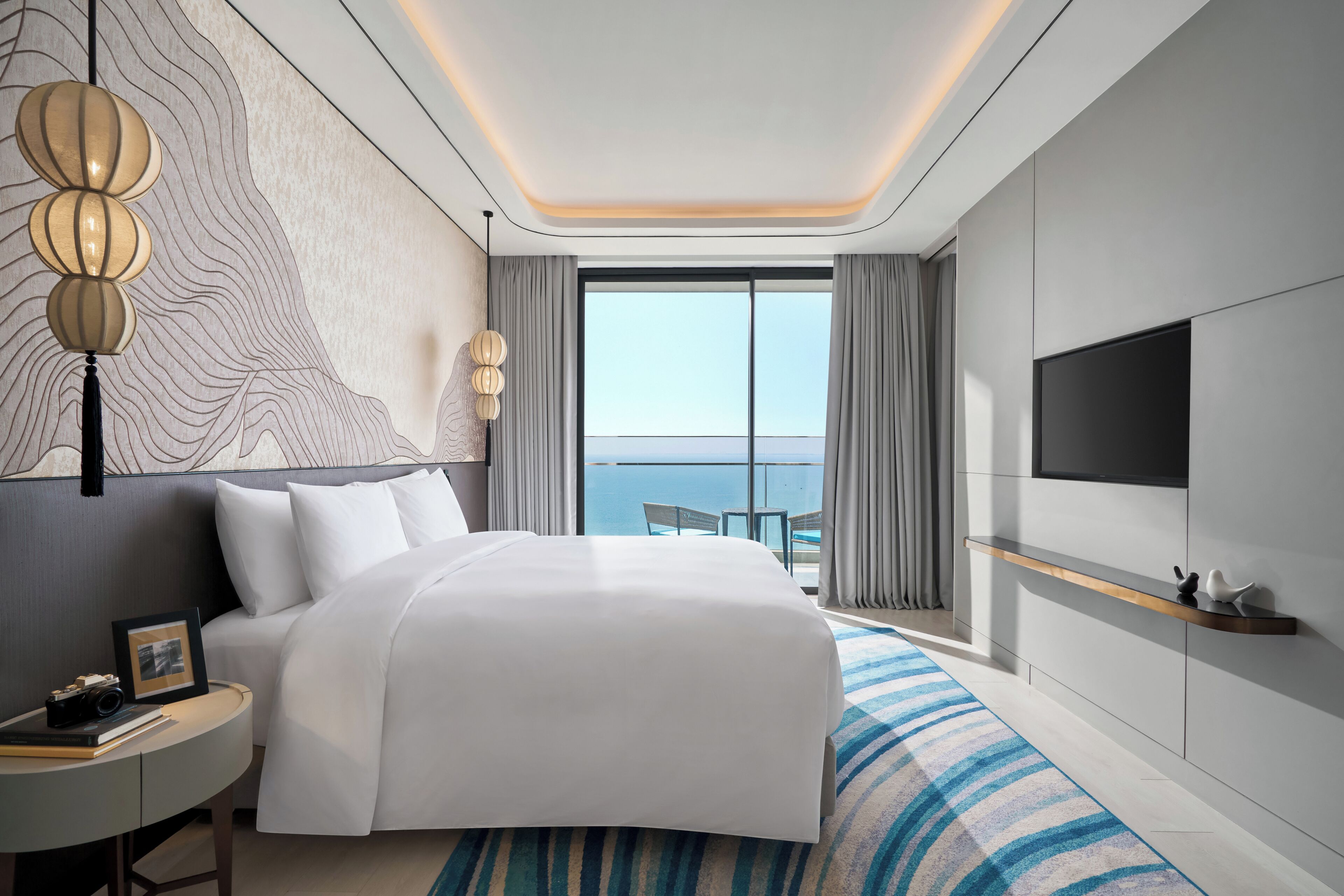 A modern hotel room with a large bed, a TV mounted on the wall, and a view of the sea through the window.