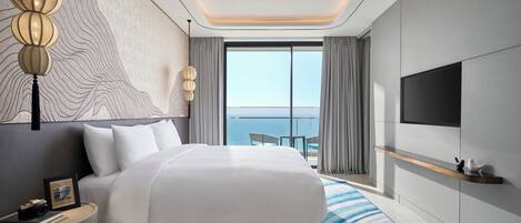 A modern hotel room with a large bed, a TV mounted on the wall, and a view of the sea through the window.