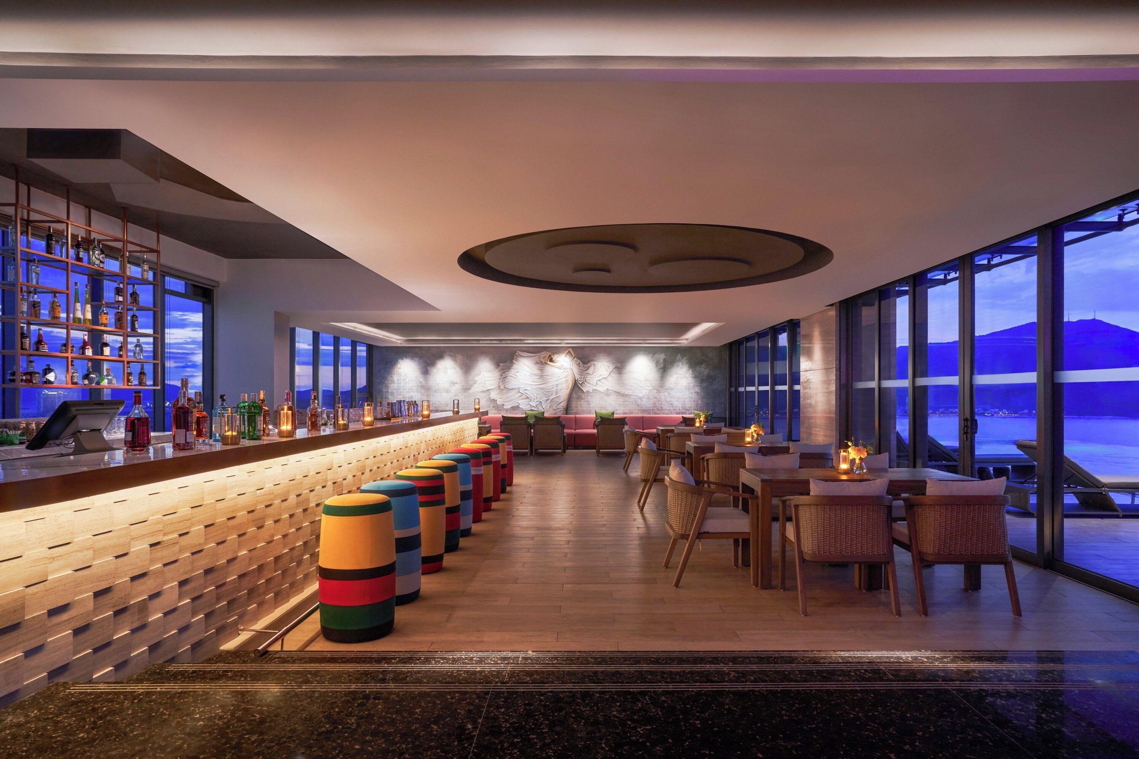A bar area with a long counter, colorful stools, and a view of the sea through large windows.