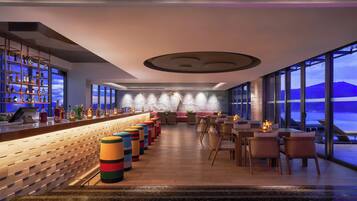 A bar area with a long counter, colorful stools, and a view of the sea through large windows.