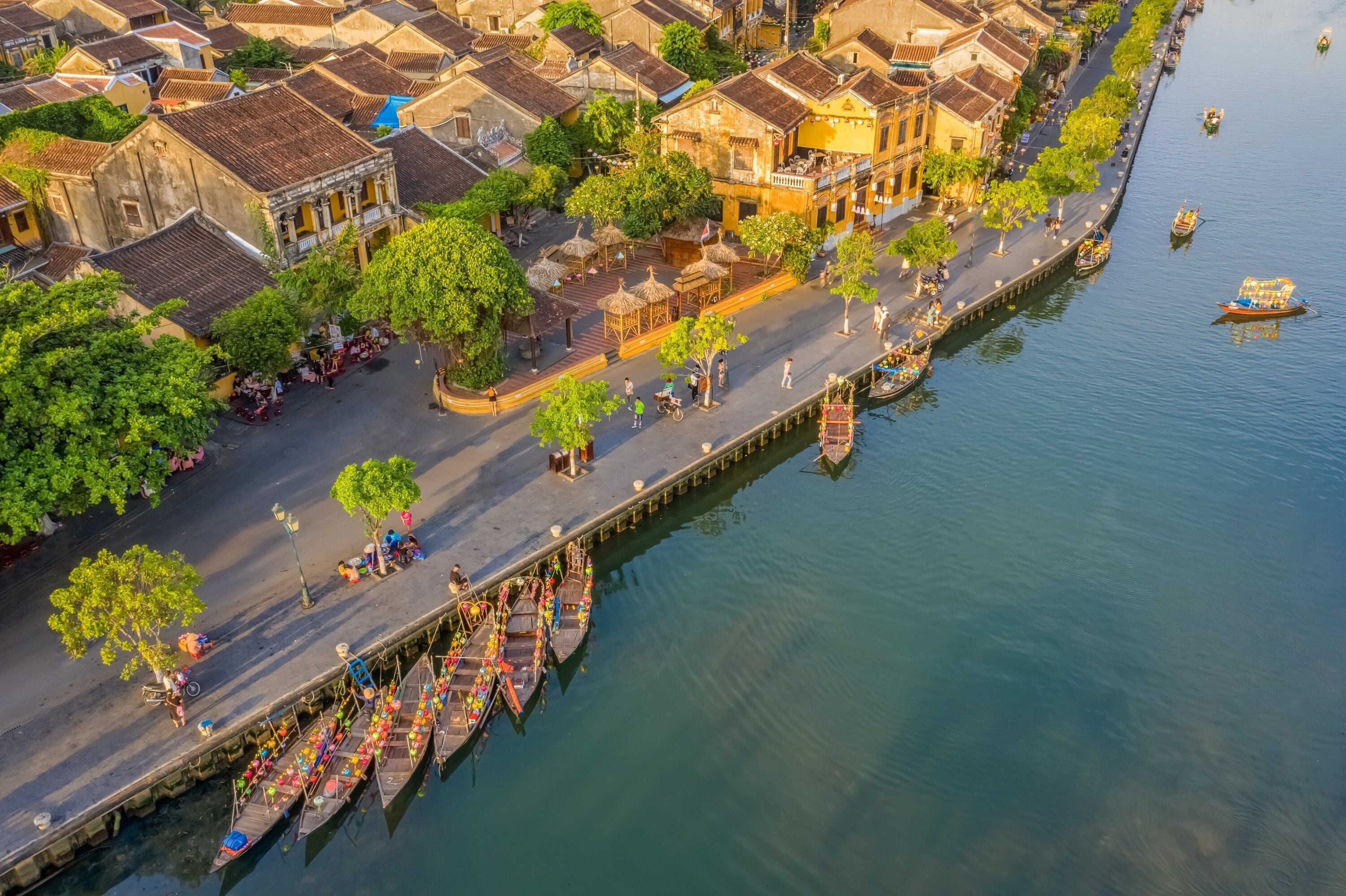 An aerial view of a riverside town with traditional buildings, a dock with boats, and a pedestrian walkway.