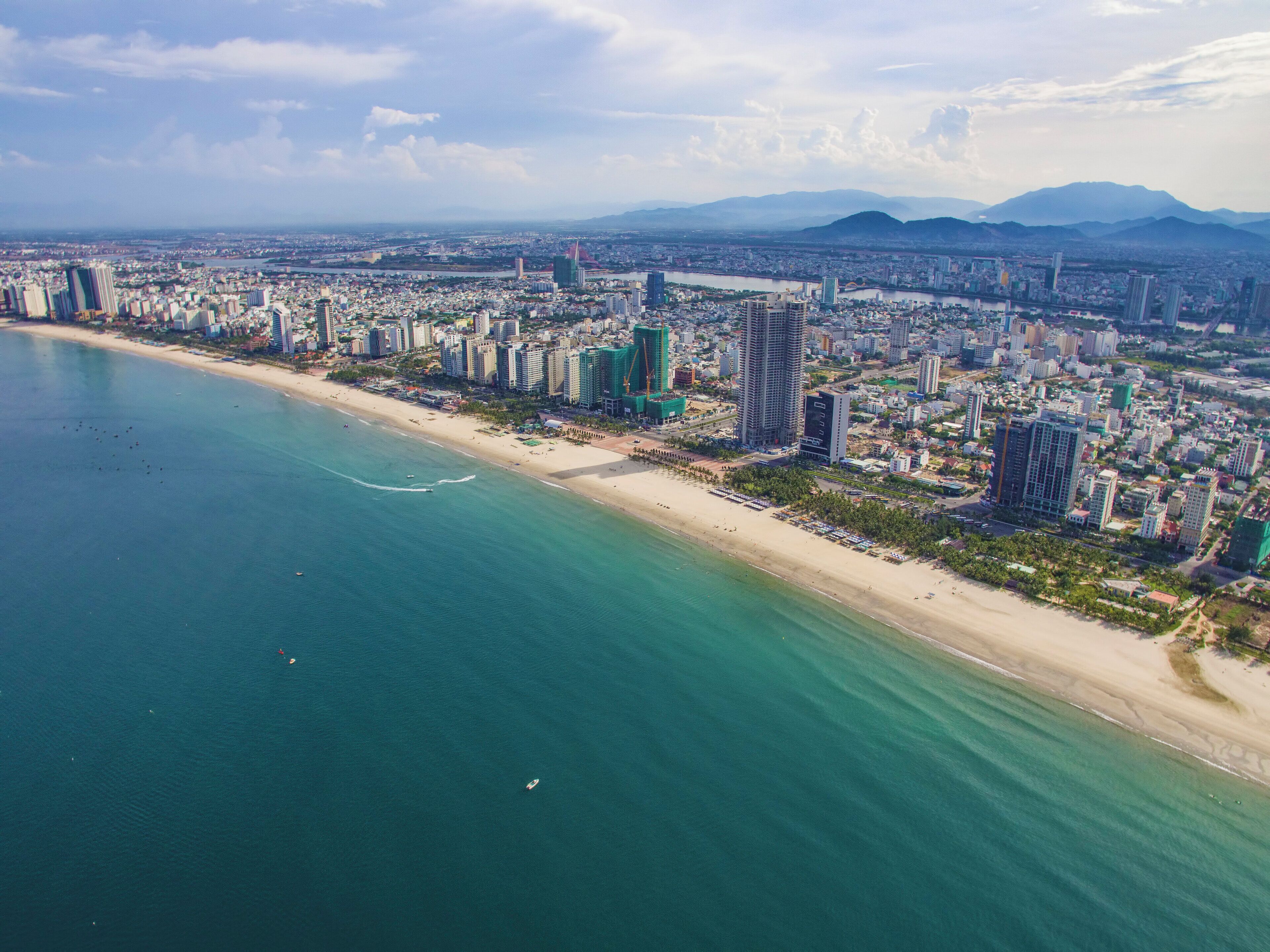 An aerial view of a coastal city with high-rise buildings, a beach, and mountains in the background.
